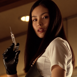 Asami Yamazaki in a white blouse, holding a syringe in a black-gloved hand